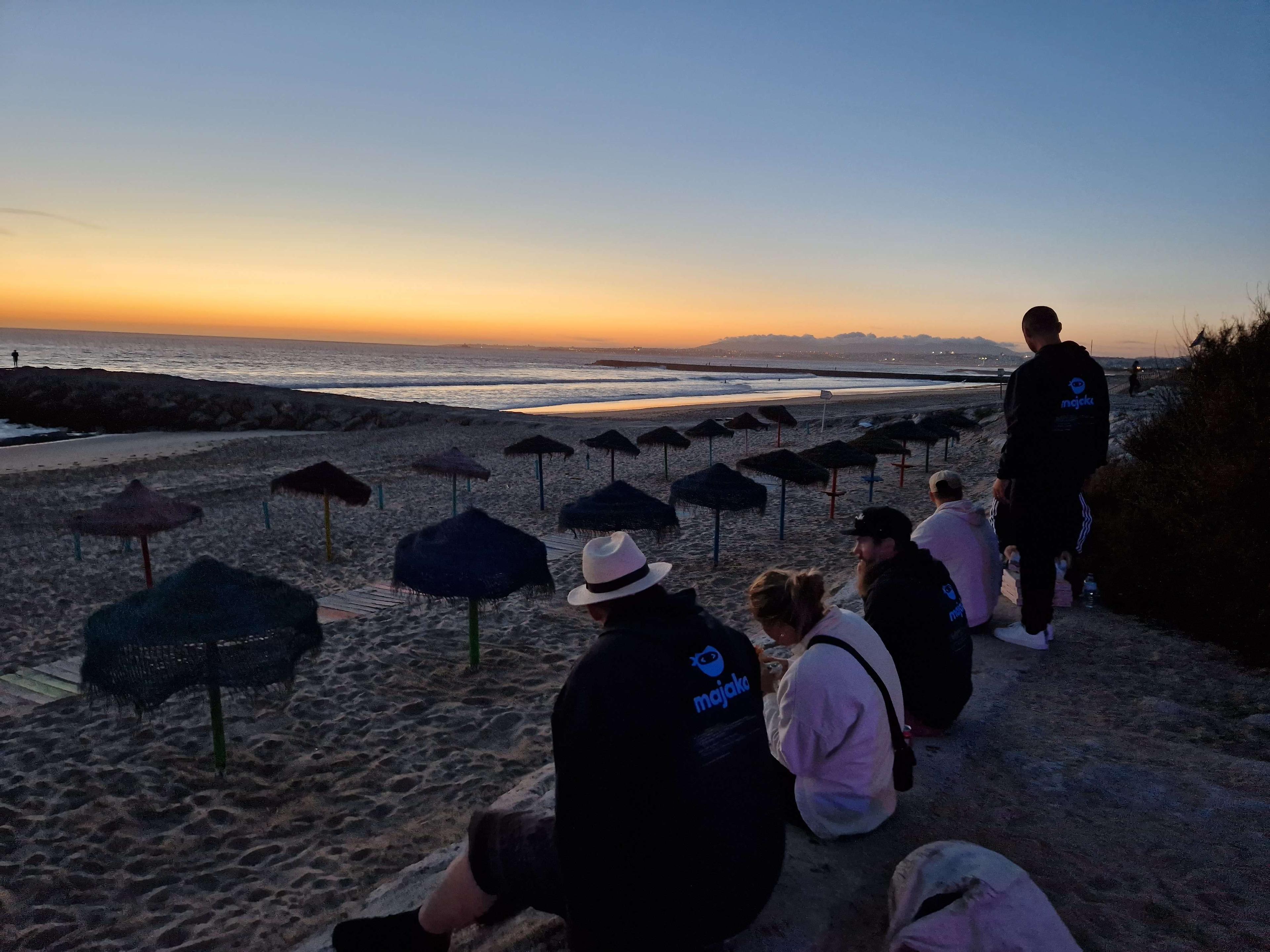 Tre personer sitter ned och två står upp vid havet, de tittar på solnedgången.