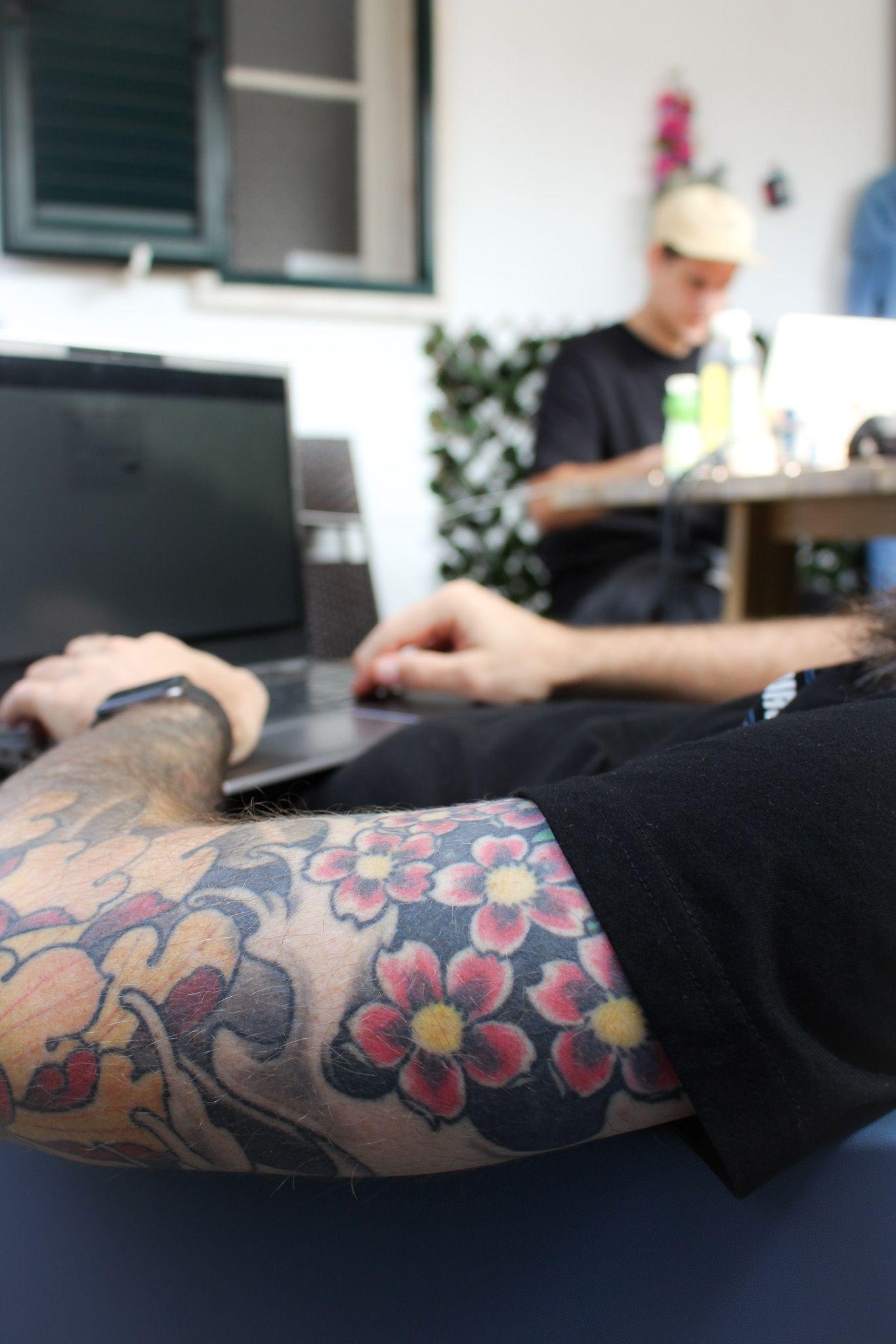 Tatuerad arm på en man som arbetar vid sin dator. I bakgrunden sitter en kollega vid ett bord.